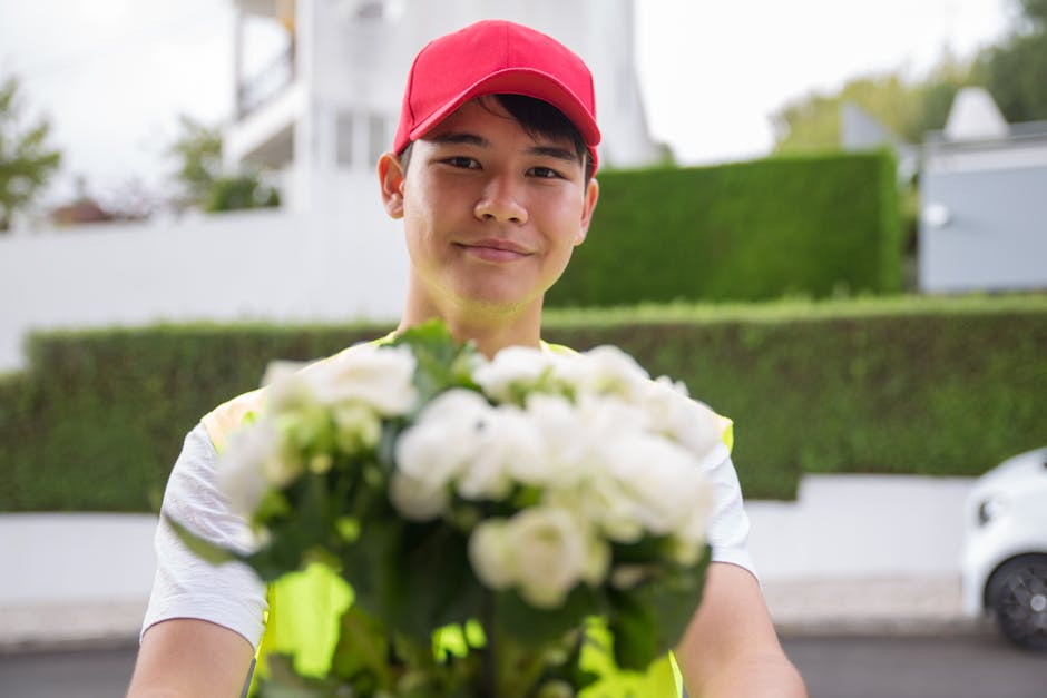 A delivery person wearing a red cap and a high-visibility yellow vest is holding out a bouquet of fresh white roses and lilies, arranged with green foliage, towards the camera. The bouquet is wrapped 
