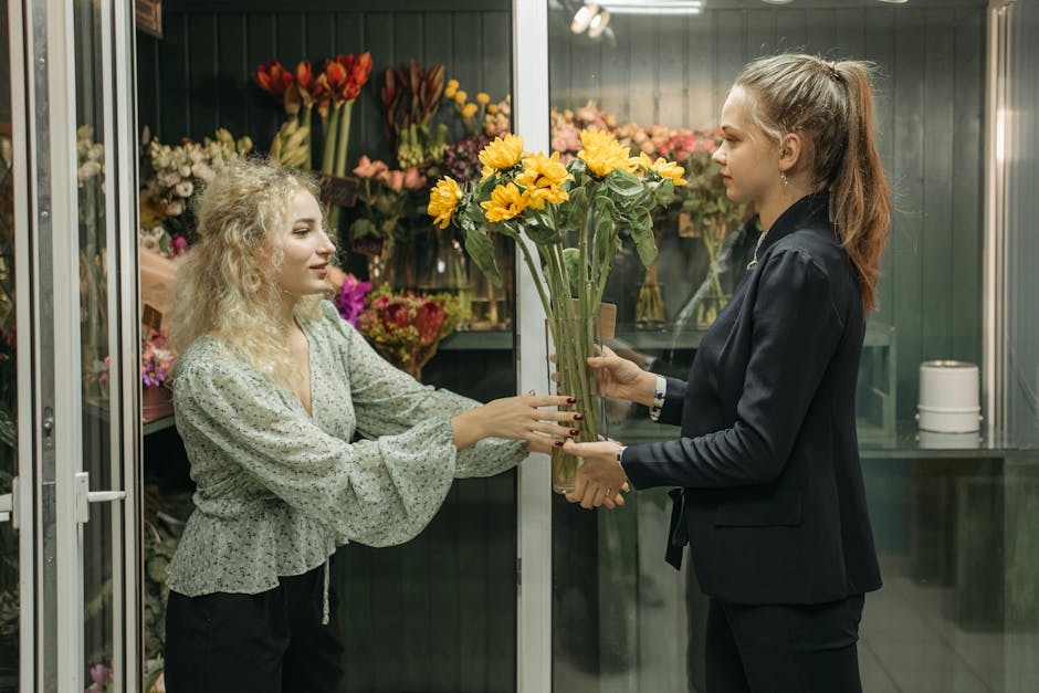 A floral arrangement consisting of bright yellow sunflowers being handed over from a woman with blonde, curly hair wearing a green floral blouse to another woman with straight, light brown hair tied b