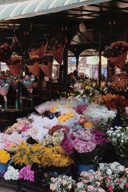 A vibrant flower stall at an outdoor market displays a diverse array of fresh floral arrangements. Brightly colored flowers, including pink, white, yellow, and purple blooms, are organized in buckets 