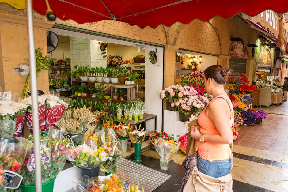 A woman browsing a flower shop with an array of colorful fresh flowers and floral arrangements displayed on tables and shelves. The flowers include pink roses, purple chrysanthemums, and mixed bouquet