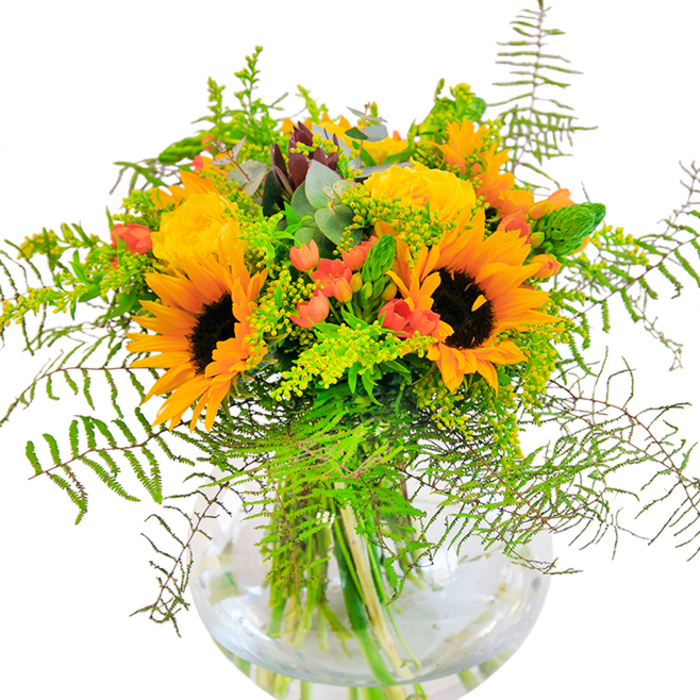 At first glance, this vibrant arrangement crafted in the heart of Poplar is pure sunshine captured in glass. Towering sunflowers preside over the bouquet, their layered yellow-orange petals opening widely around rich brown centers-a tribute to the exuberance of late summer afternoons by Limehouse Cut. Interwoven are velvety yellow garden roses, their soft spirals lending a gentle formality and subtle, inviting fragrance. Dotted throughout, clusters of red Hypericum berries glimmer with a cherry-bright sheen, contrasting playfully against drifts of lush, deeply green foliage: feathery ferns, slender spikes, and light green filler, perhaps goldenrod, speckled with minuscule yellow blossoms. The bouquet's shape is natural yet refined, its composition brought together in a flawless, rounded glass vase that lays bare the vibrant stems within, magnified by the water's pristine clarity. Bluish eucalyptus leaves, with their silvery touch, add a rustic accent and complete the sense of abundance. The arrangement glows in the soft, ambient Poplar light, reminding the viewer of a peaceful garden afternoon. Thoughtful and expressive, this bouquet is suited for a host of special occasions-a birthday, an anniversary, or even to light up a quiet workspace. Its fresh scent and bright palette evoke optimism and comfort, reflecting the charm and warmth found throughout the local E14 neighborhoods and bustling markets.