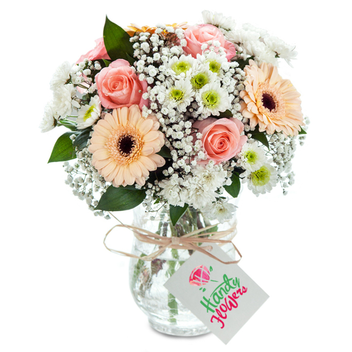 Arranged against a glowing white background, this expressive bouquet is the work of masterful florists in Poplar, reflecting the area's blend of tradition and vibrancy. Plush peach-pink roses, their petals elegantly layered and softly curled, form a romantic centre, exuding both fragrance and warmth. Two prominent gerbera daisies, rendered in gentle peach tones with signature dark centers, radiate brightness that echoes the inviting atmosphere of Poplar's Sunday flower stalls. Baby's breath, white and cloudlike, fills the spaces between, lending an ethereal, lofty feel suggestive of open skies above the docks. White chrysanthemums with flashes of lime at their center add textural complexity and youthful optimism to the bouquet, complemented by sprigs of eucalyptus whose subtle green shades conjure memories of parkland strolls along the River Lea. All the elements are gathered into a clear, textured glass vase, revealing clean stems and sparkling water. The arrangement is tastefully finished with a bow of natural raffia and a blank tag-ready for personal sentiments. This bouquet is a versatile expression of care, ideal for birthdays, tributes, or everyday gestures, and is available for quick delivery across Poplar.